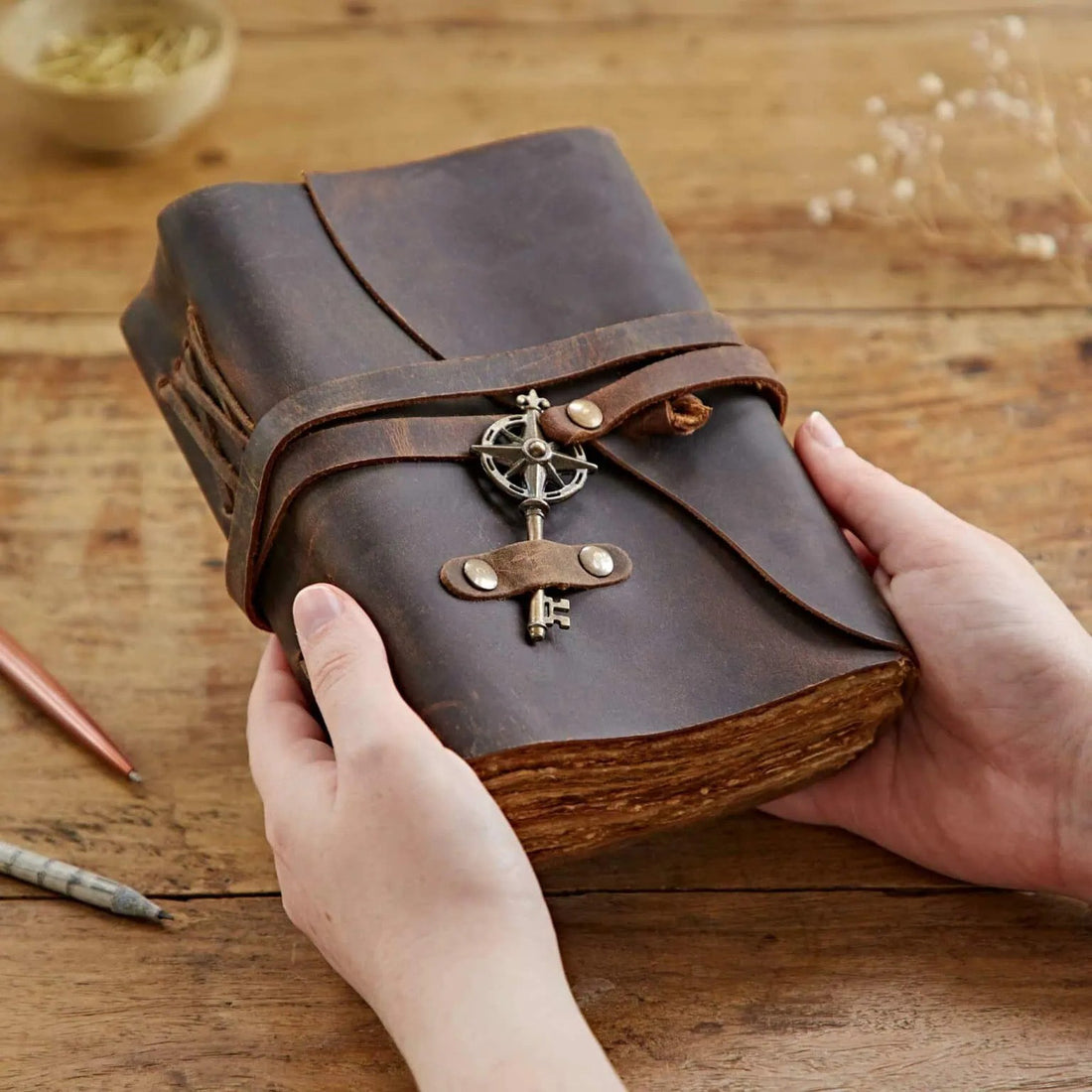 A hand holding an open leather journal with a metal compass key and a leather strap, on a wooden surface.