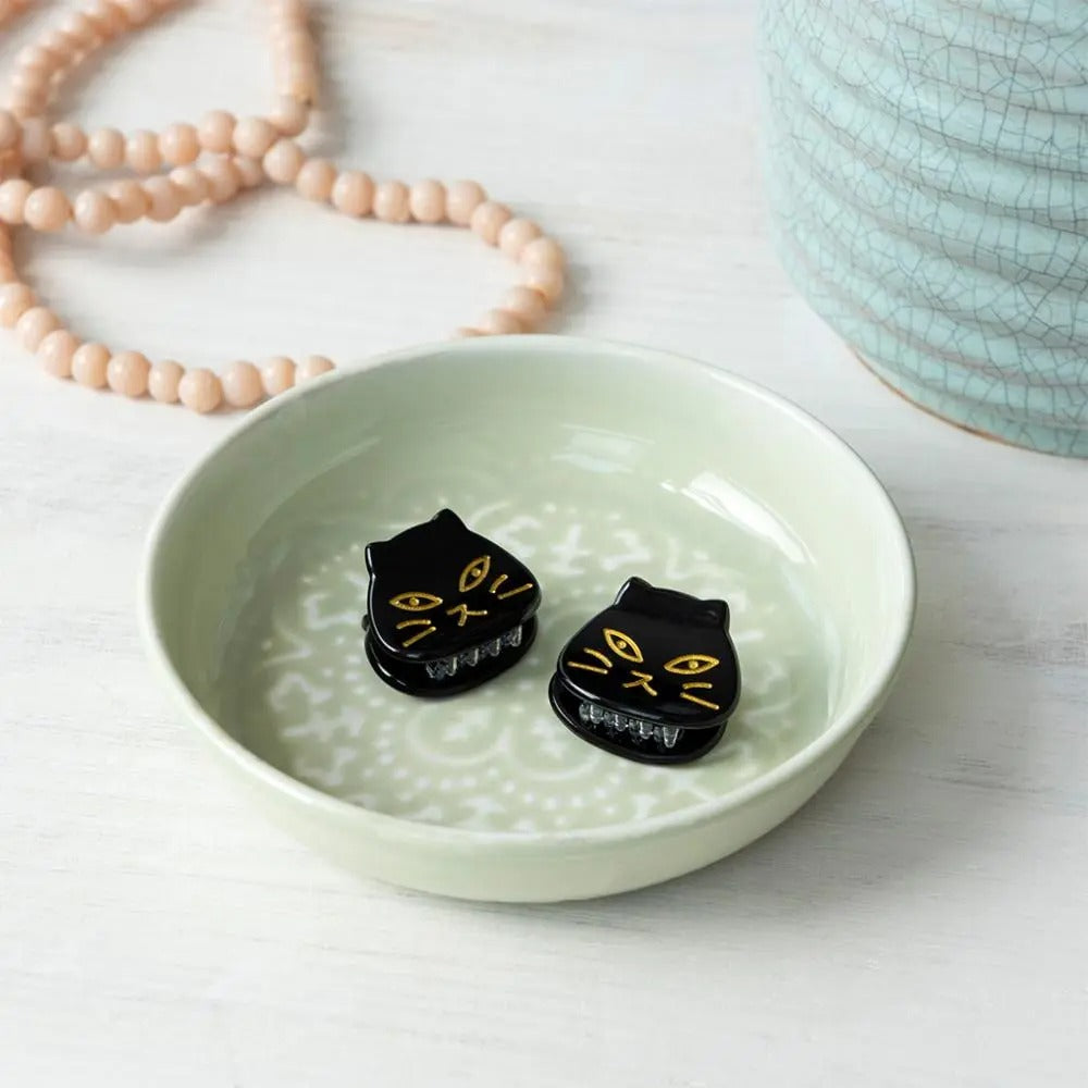 Black cat-shaped earrings on a light green ceramic dish with a necklace and ceramic item in the background.
