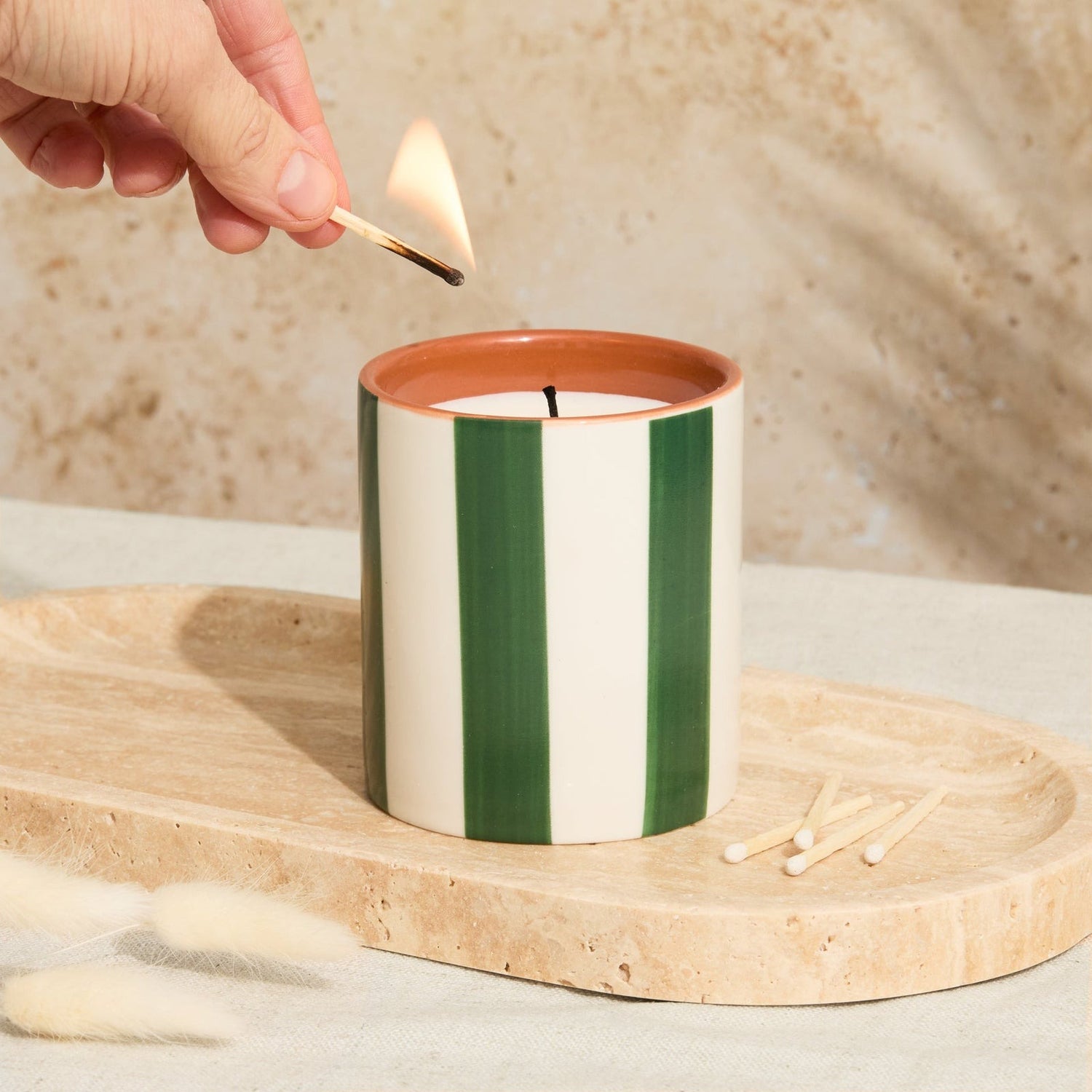 A person lighting the Olive Stripe Candle in ceramic vessel with matching lid and oud leather fragrance