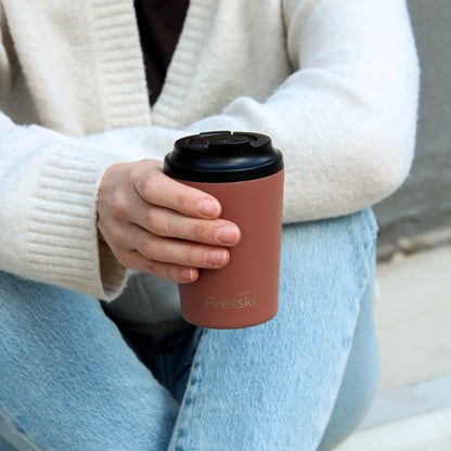 Person holding a tuscan reusable coffee cup with a black lid, wearing a beige sweater and light blue jeans.