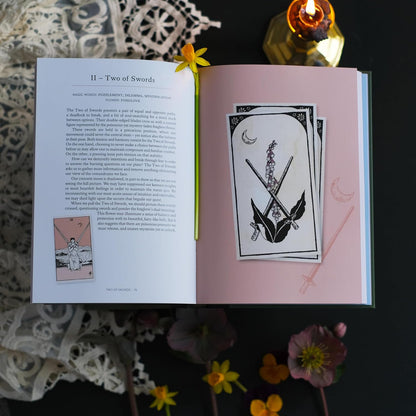 Open book with tarot card illustration of two of swords and text on a dark surface with flowers.