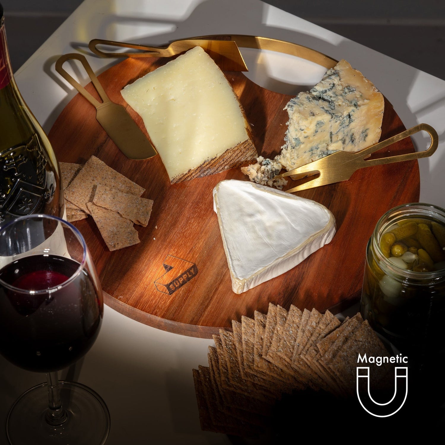 Wooden cheese board with assorted cheeses, crackers, and a glass of red wine on a white surface.