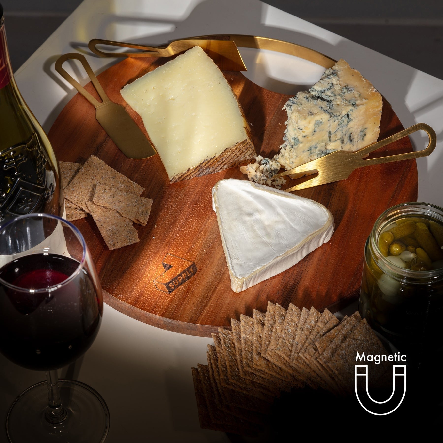 Wooden cheese board with assorted cheeses, crackers, and a glass of red wine on a white surface.