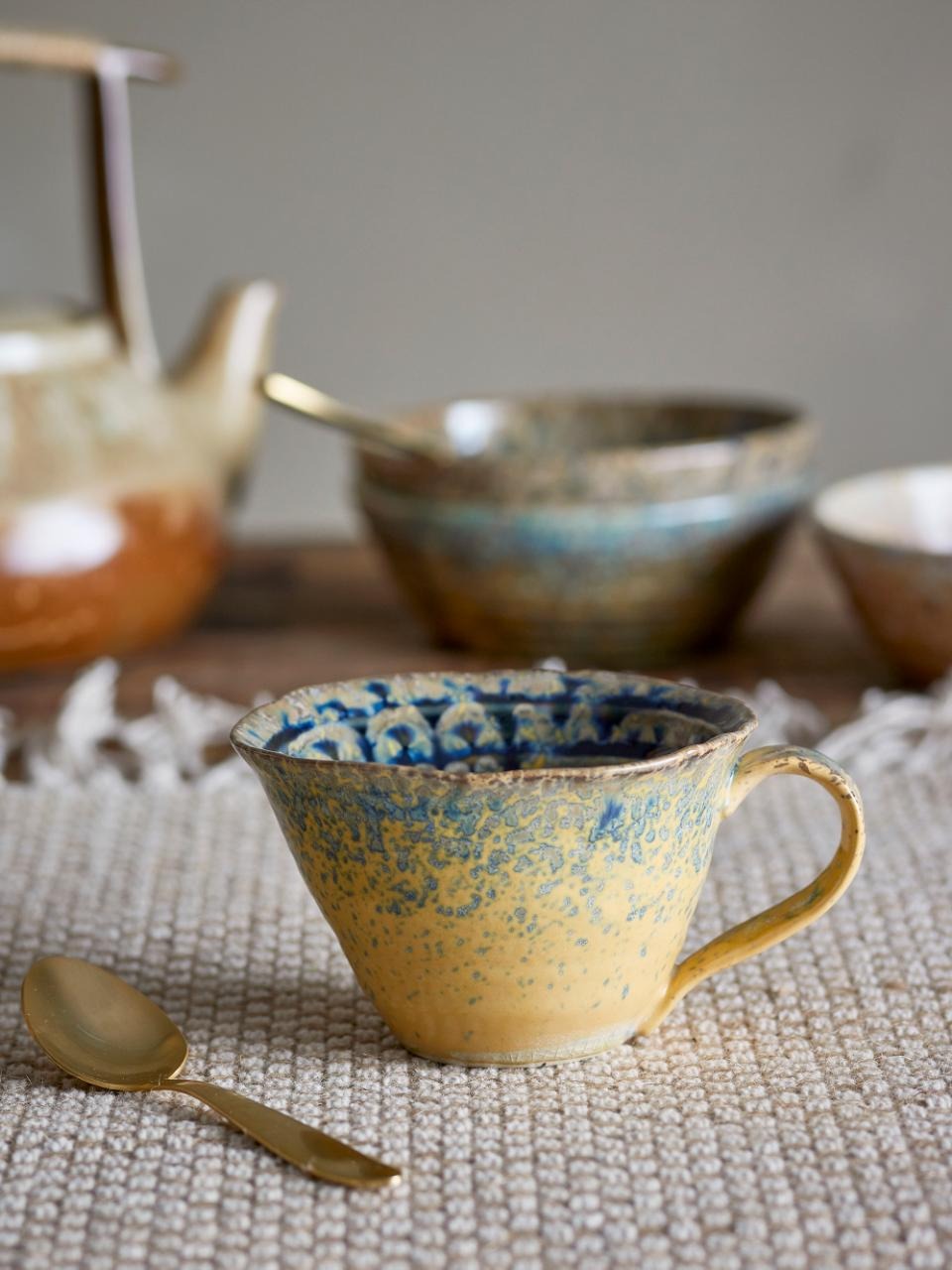 Handcrafted ceramic cup with a reactive yellow and blue glaze, featuring pearlescent shimmer highlights, placed on a rustic wooden table with a gold spoon beside it.