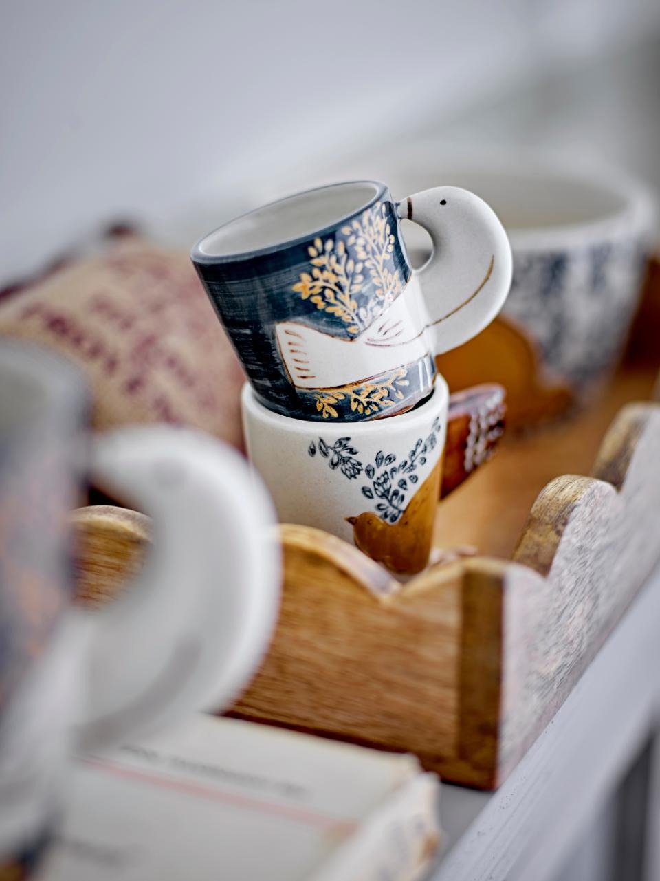 Two Hezha espresso cups stacked on top of each other inside a wooden tray, one in blue and one in nature color, each featuring a delicate bird motif with wings as handles. Hand-decorated ceramic cups, unique and ideal for espresso or display.